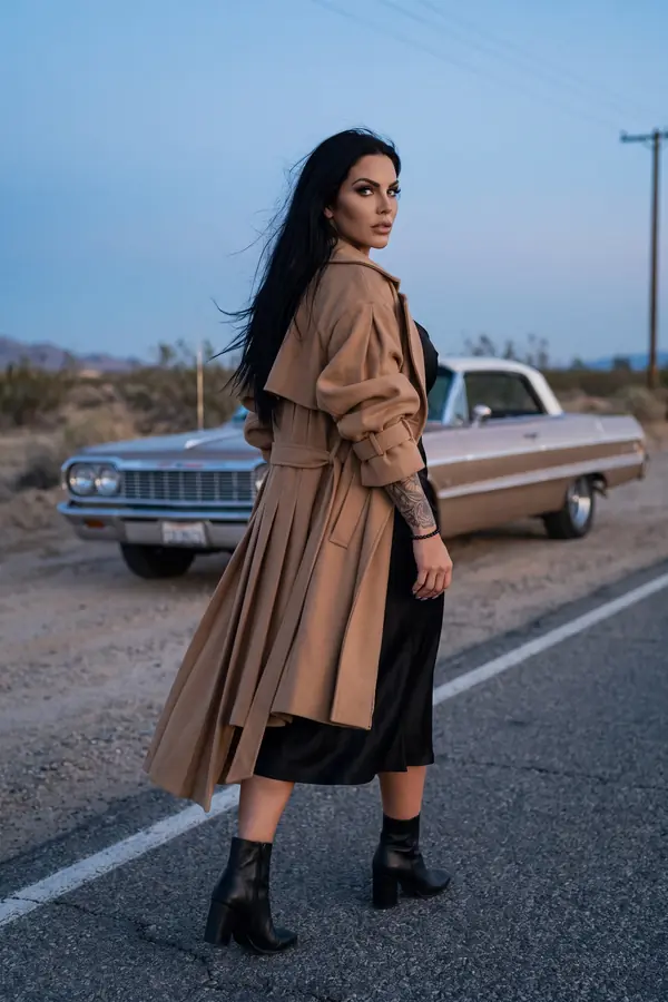 Woman seen from behind in a long camel wool trench over a black satin slip dress on a desert road at dusk