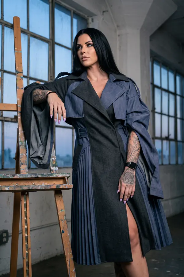 Woman in a charcoal wool and blue denim trench dress with pleated panels and draped sleeves leaning on an easel in a loft