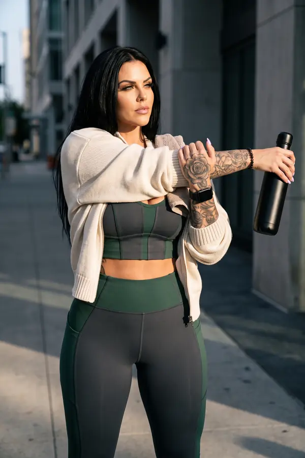 Woman in a charcoal and green colour-block sports top and leggings with a cream knit cardigan, stretching on a city sidewalk