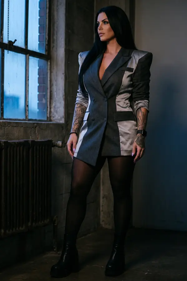 Woman in a charcoal and silver colour-block blazer dress with black tights and platform ankle boots by an industrial window