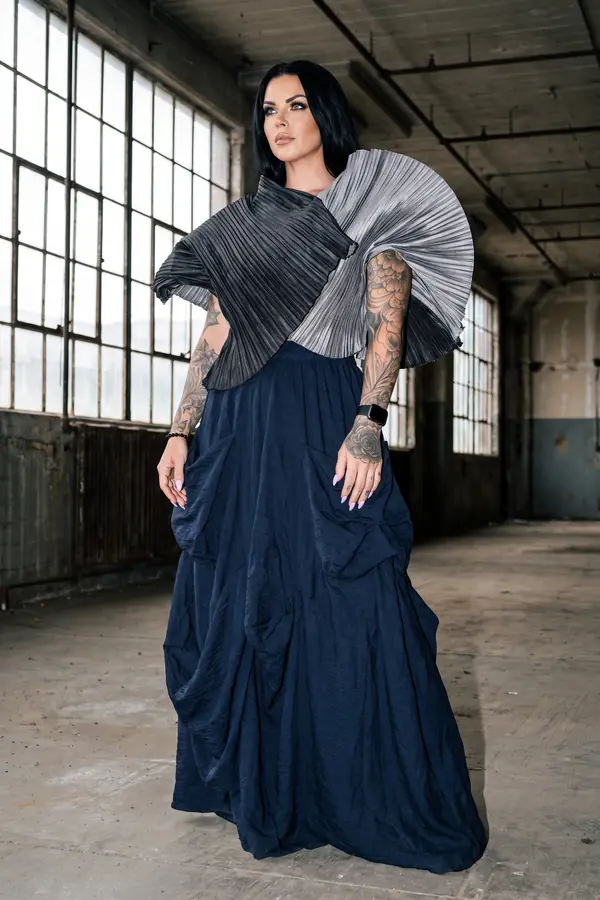 Woman in a knife-pleated charcoal and silver fan top and a navy crinkle maxi skirt in an empty warehouse