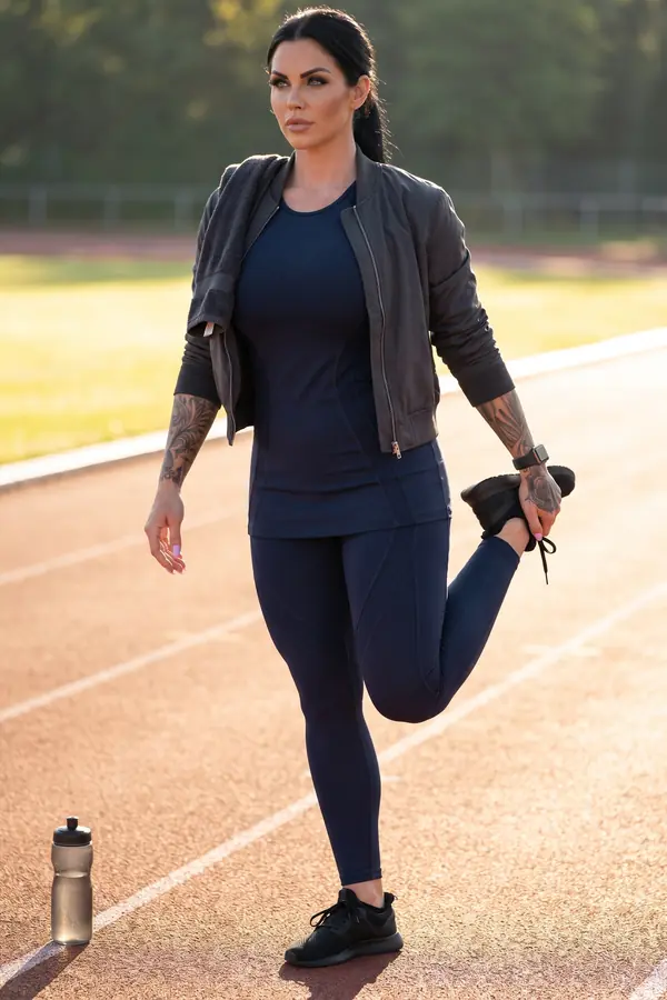 Woman in a navy fitted top and leggings with a charcoal bomber, stretching one leg behind her on a red running track