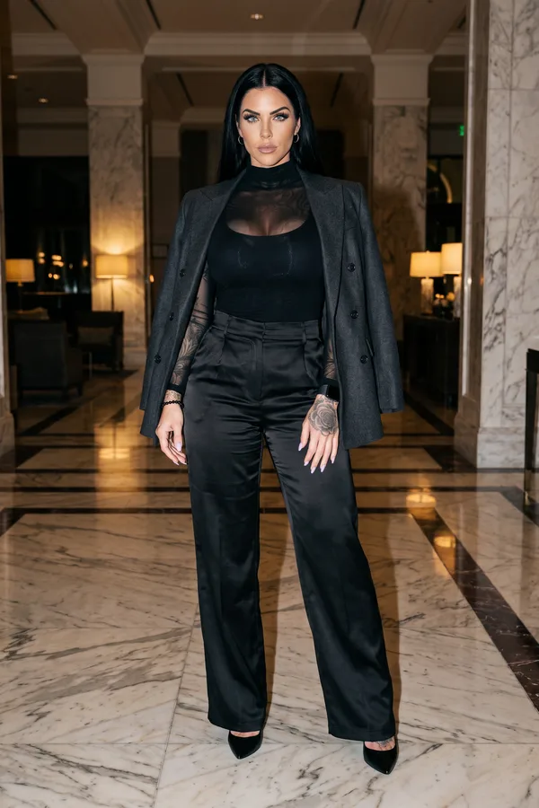Woman in a charcoal double-breasted blazer, black mesh turtleneck and black satin wide-leg trousers in a hotel lobby