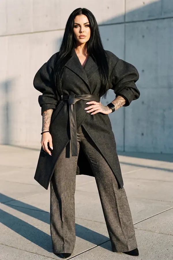 Woman in a charcoal balloon-sleeve coat with a wide leather obi belt and herringbone wide-leg trousers by a concrete wall