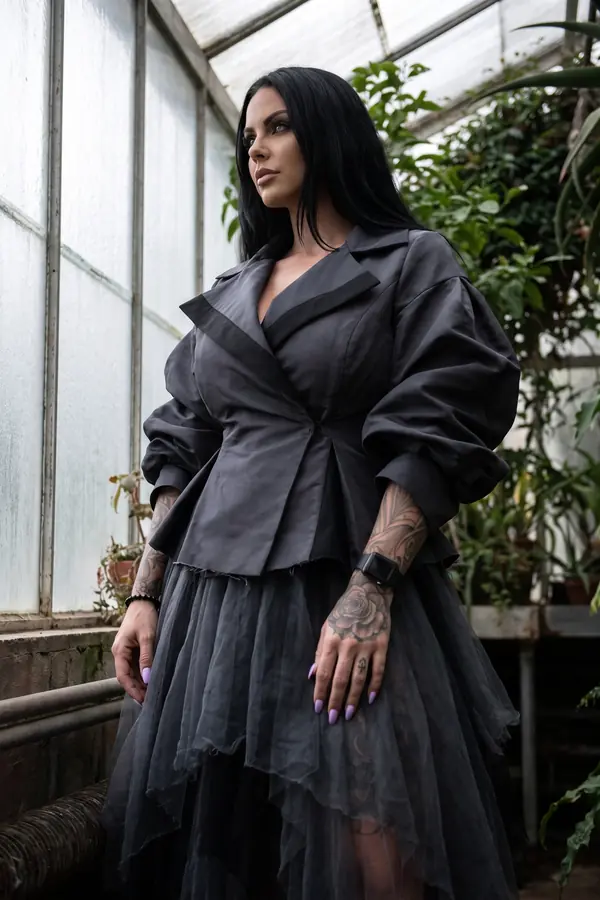 Woman in a charcoal taffeta jacket with puff sleeves and a tiered grey tulle skirt standing in a greenhouse