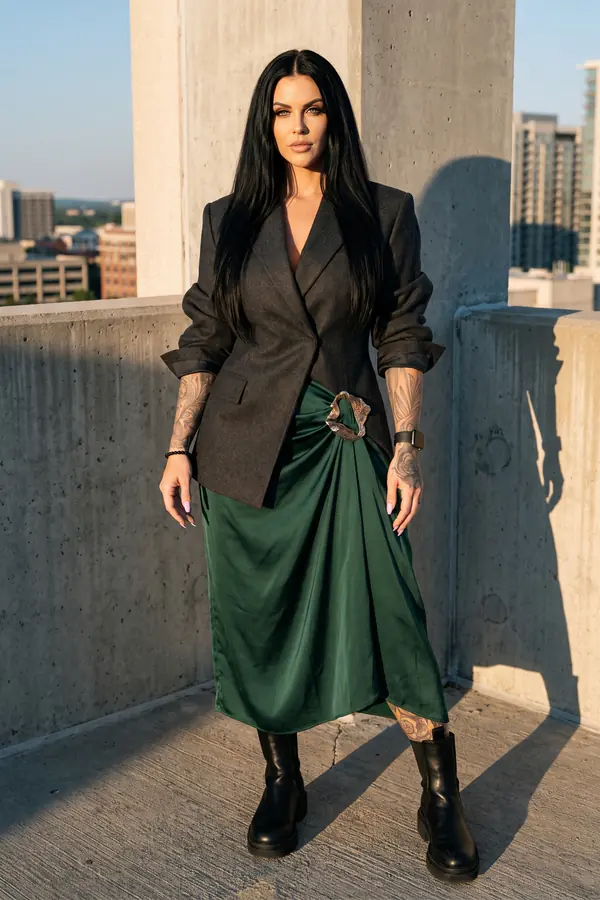 Woman in an oversized charcoal wool blazer and emerald satin midi skirt with a gold buckle on a parking rooftop at sunset