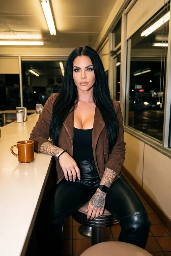 Woman seated at a diner counter in an open chocolate suede jacket, black tank and black leather trousers with a coffee mug