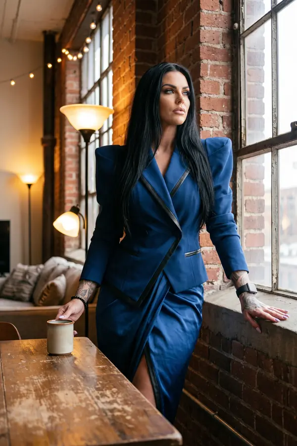 Woman at a loft window in a cobalt blazer with black leather trim and a cobalt satin wrap skirt, coffee mug on the table