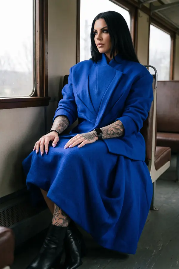 Woman in a cobalt boiled-wool jacket and draped midi skirt seated by a train window with black ankle boots