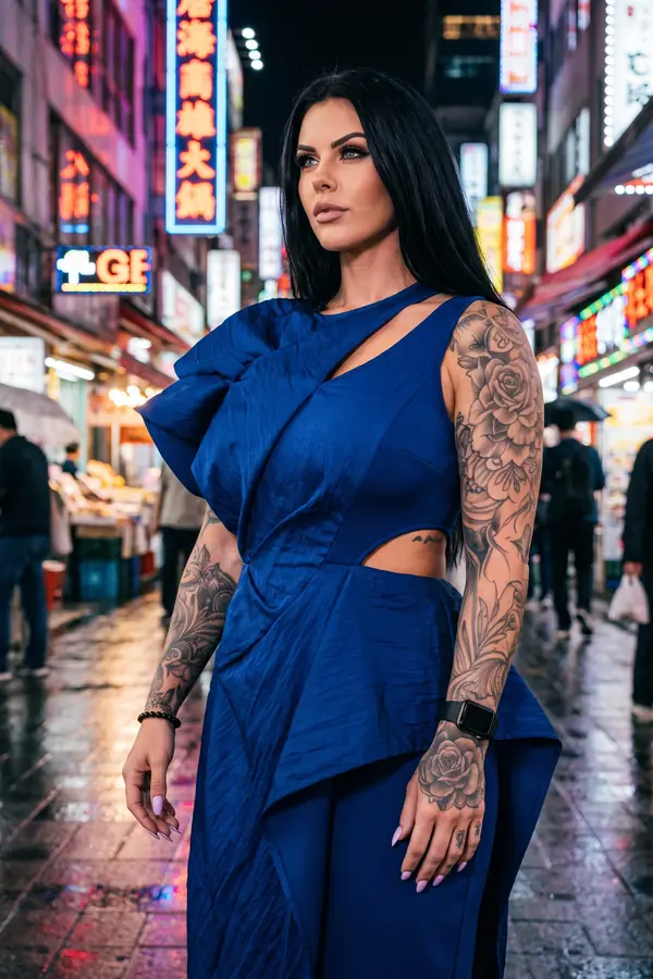 Woman in a cobalt crinkled satin draped dress with a shoulder ruffle and waist cut-out on a rainy neon-lit street