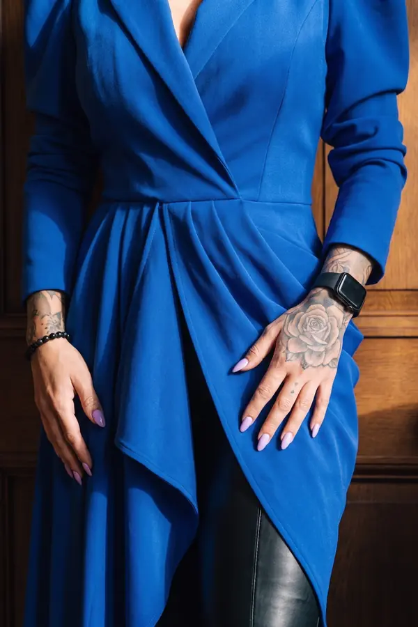 Woman in a cobalt draped blazer dress with padded shoulders and black leather trousers in a wood-panelled hotel lounge
