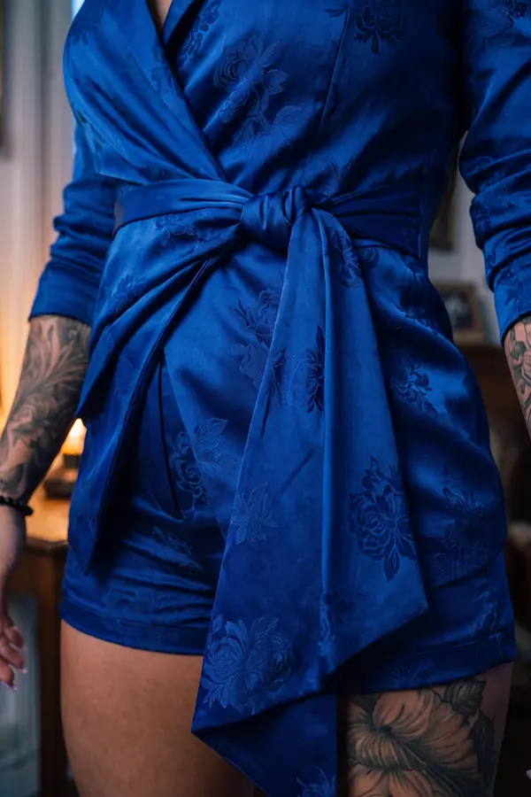 Woman in a cobalt floral jacquard satin wrap blazer tied with a sash and matching shorts by a tall window at dusk