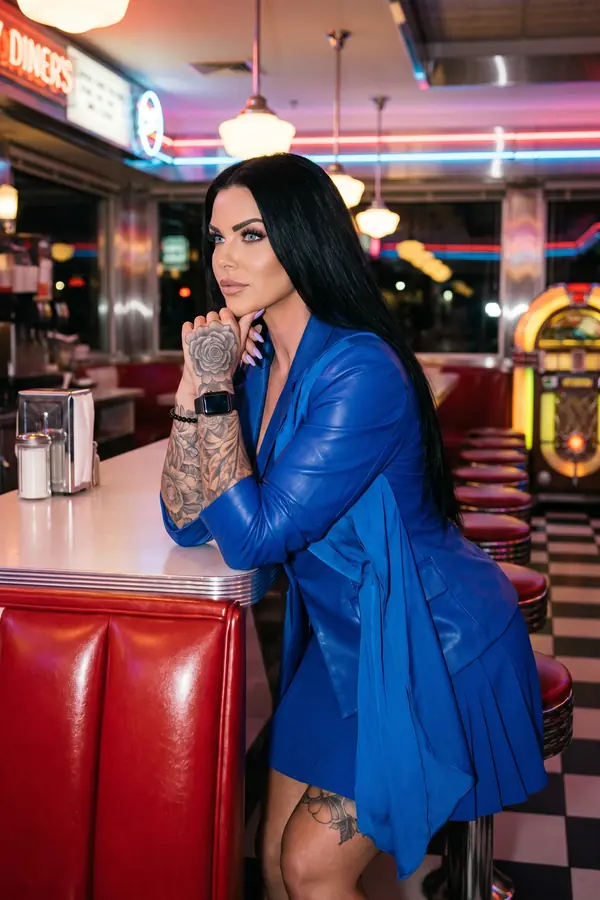 Woman in a long cobalt leather blazer with a satin drape and a knife-pleated mini skirt, leaning on a diner counter at night