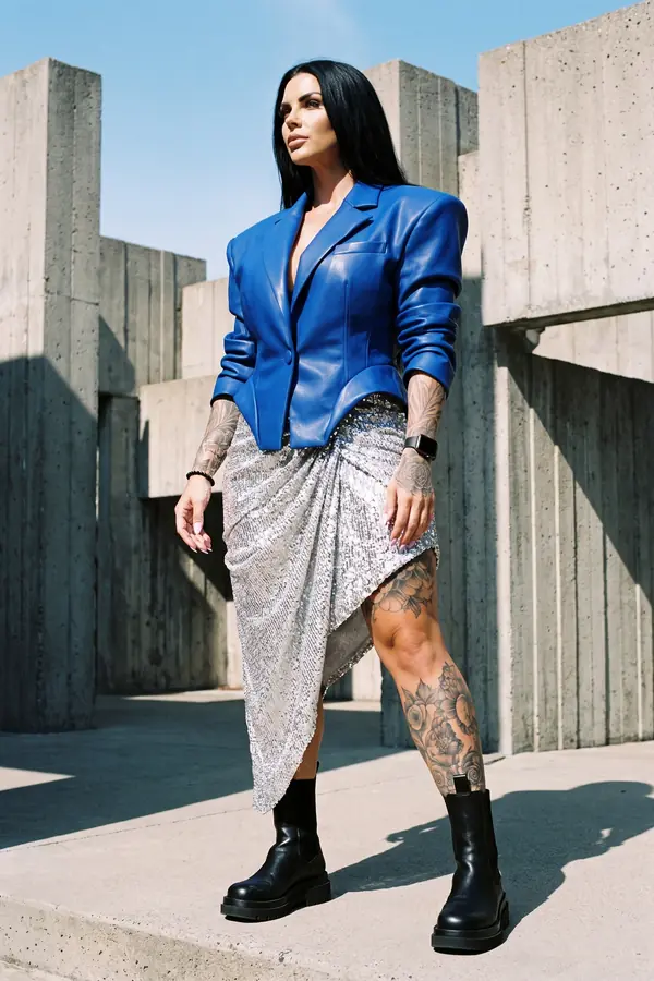 Woman in a fitted cobalt leather blazer and silver sequin draped skirt with black chelsea boots against a concrete wall