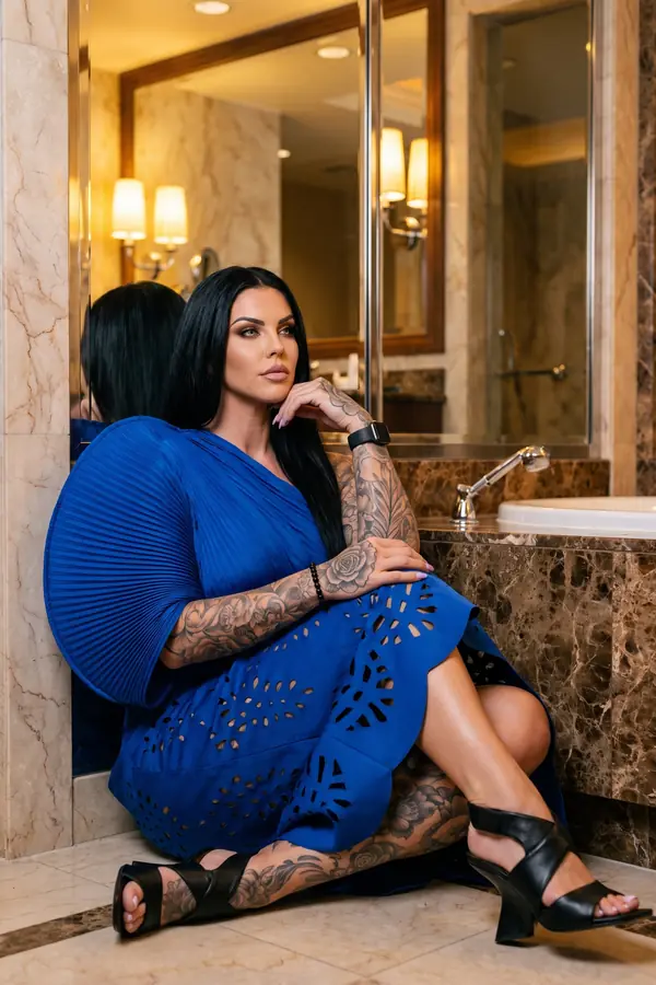 Woman in a cobalt pleated dress with a laser-cut skirt and black sandals sitting on a marble hotel bathroom floor