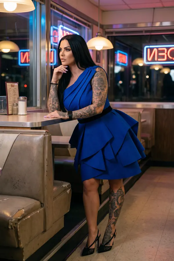 Woman in a cobalt one-shoulder mini dress with a tiered origami skirt and black velvet waistband, leaning on a diner table