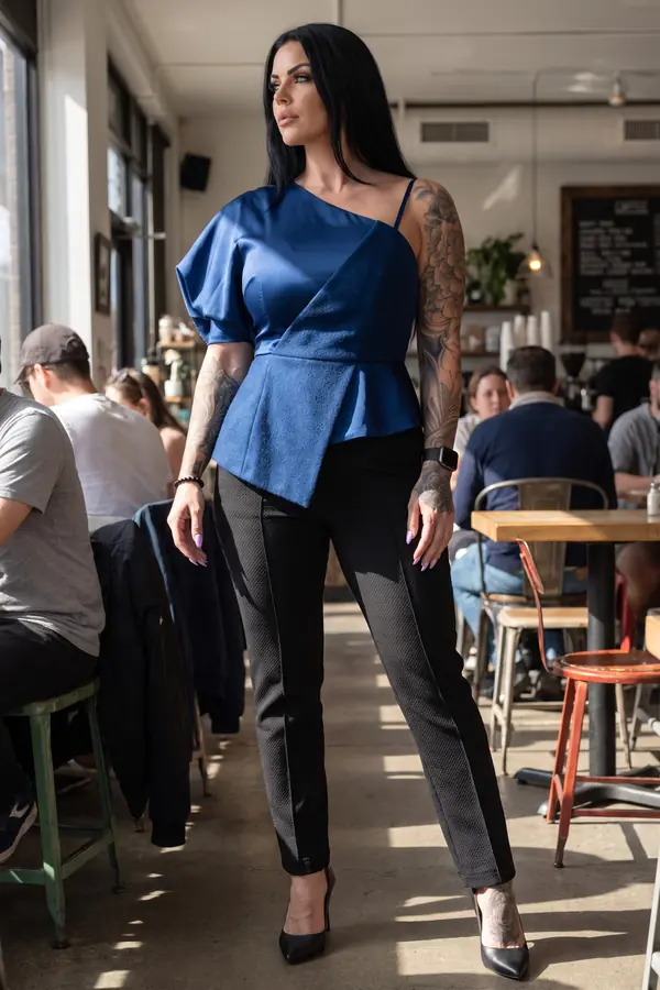 Woman in a cobalt one-shoulder satin and wool peplum top with black slim trousers and pumps in a sunny coffee shop