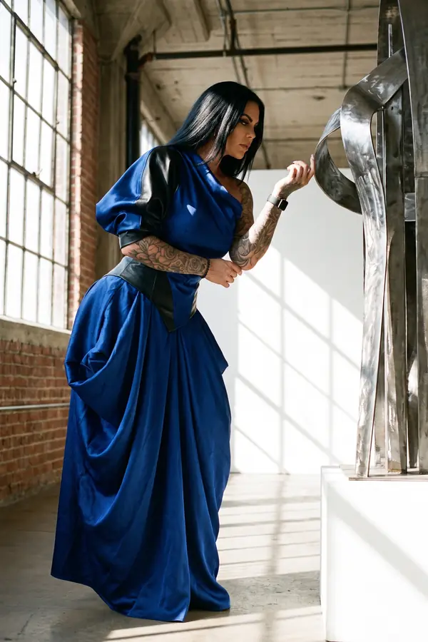 Woman with long black hair in a cobalt one-shoulder satin gown with black leather panels leaning towards a steel sculpture