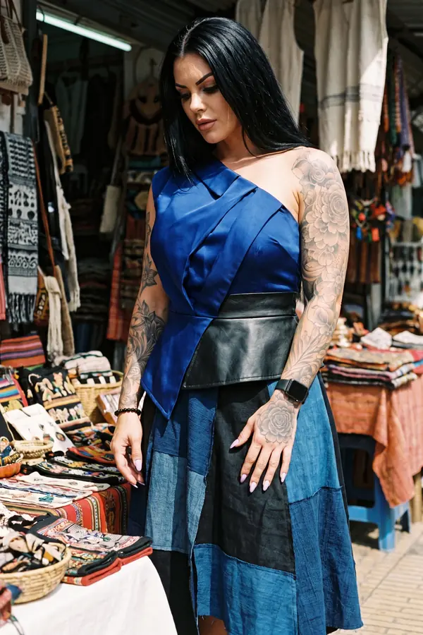 Woman in a cobalt one-shoulder satin top with a black leather waistband and a patchwork denim midi skirt at a market stall
