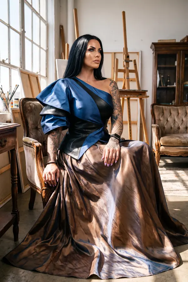 Woman seated in an artist's studio in a cobalt satin and leather one-shoulder top and a long bronze satin skirt