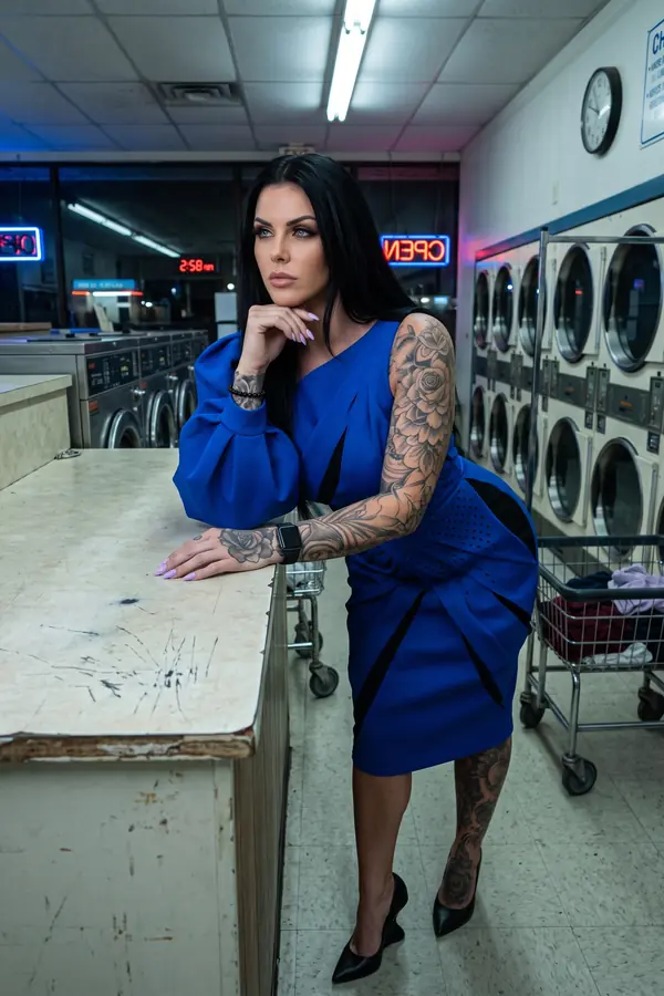 Woman in a cobalt one-sleeve midi dress with black slash panels and black pointed pumps, leaning on a laundromat counter