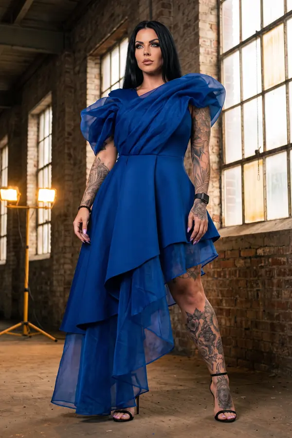 Woman in a cobalt high-low dress with organza puff shoulders and black sandals in a brick warehouse