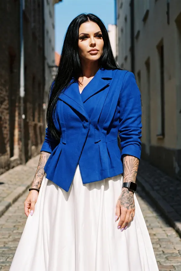 Woman in a cobalt wool peplum blazer and ivory satin circle skirt standing in a sunlit cobbled alley between old buildings