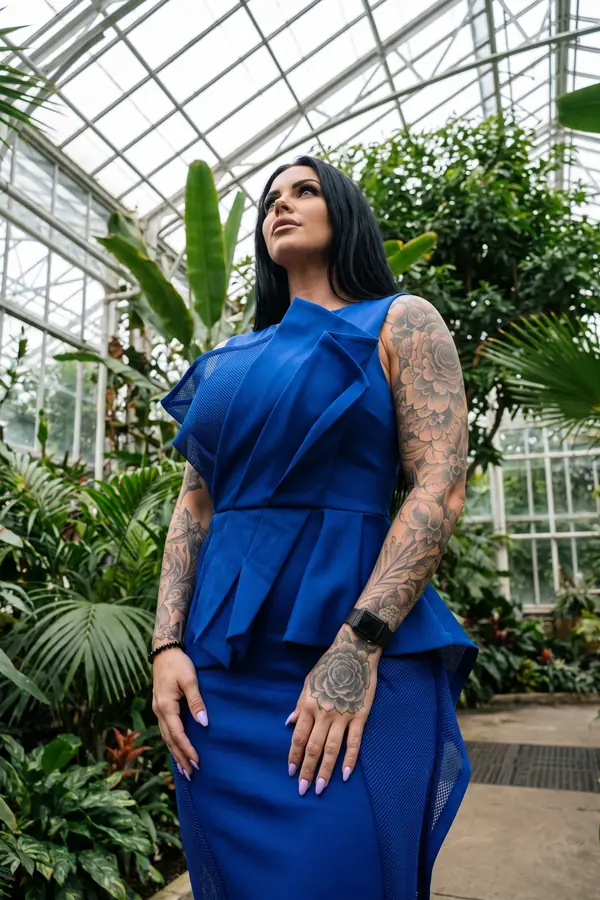 Woman in a sleeveless cobalt peplum midi dress with folded panels and mesh insets in a glass greenhouse