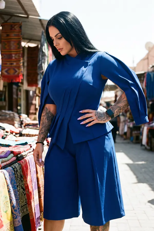 Woman in a cobalt pleated-front top with split sleeves and matching wide culottes looking down at a market stall of textiles
