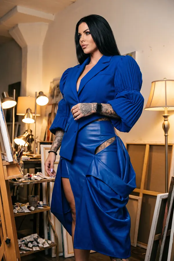 Woman in a cobalt blazer with pleated balloon sleeves and a draped cobalt faux leather skirt in a lamp-lit artist's studio
