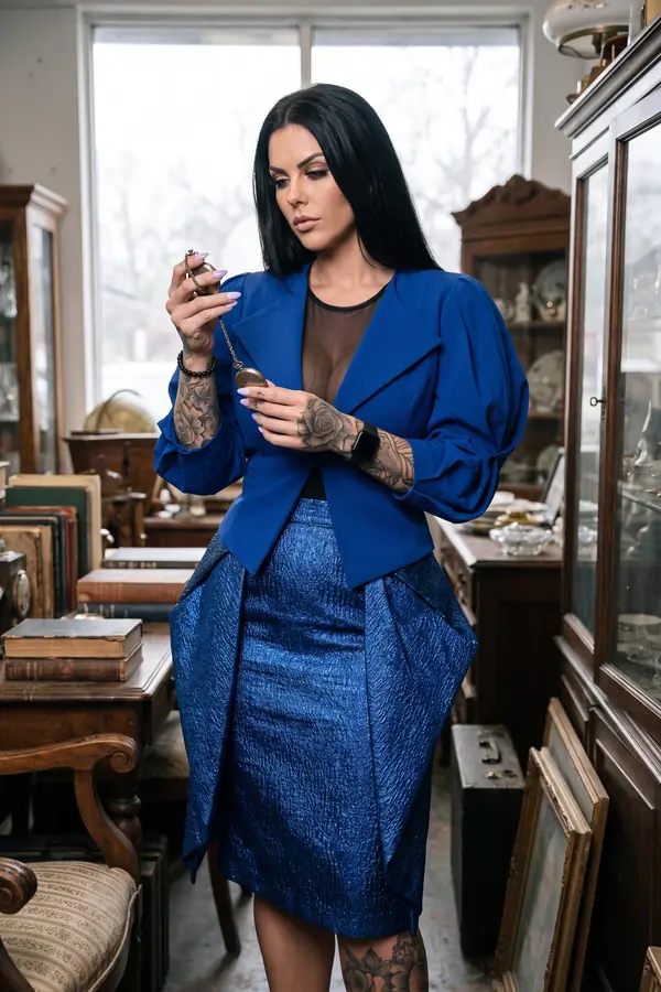 Woman in a cobalt crepe jacket with balloon sleeves, a black mesh top and a metallic blue pencil skirt in an antique shop