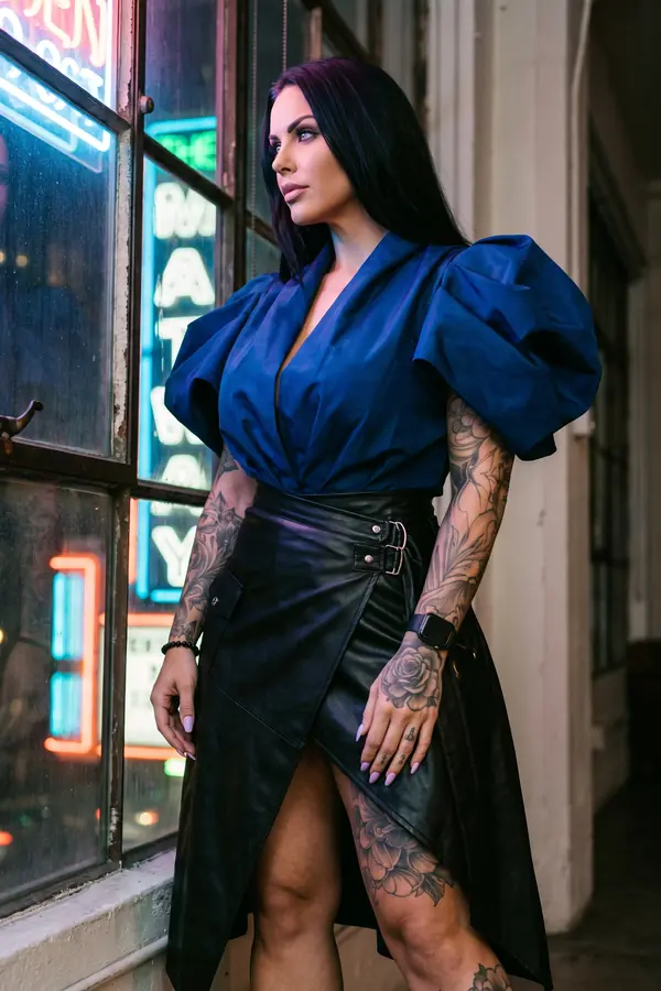 Woman in a cobalt taffeta puff-sleeve wrap top and a black leather wrap skirt with silver buckles by a neon-lit window