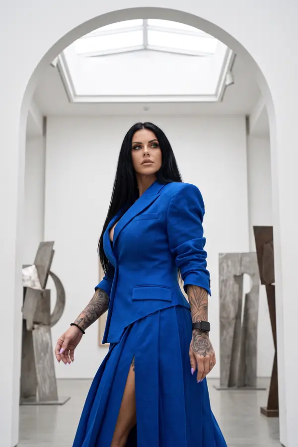 Woman in a cobalt wool blazer with a raw diagonal hem and a pleated maxi skirt walking through a white sculpture gallery