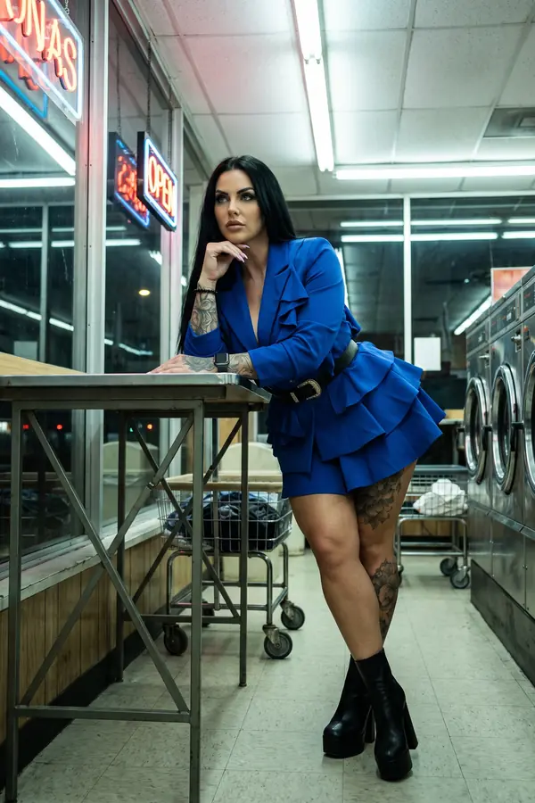 Woman in a laundromat at night in a cobalt ruffle blazer dress with a western-buckle belt and black platform boots