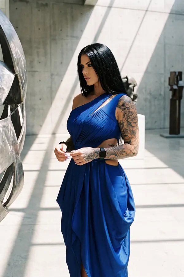 Woman in a cobalt satin one-shoulder dress with sunray pleats beside a steel sculpture in a concrete gallery