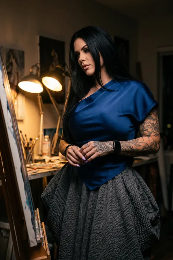 Woman in a cobalt satin asymmetric top and charcoal crinkle bubble skirt standing by an easel under studio lamps