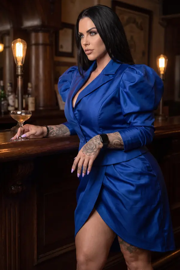 Woman in a cobalt satin blazer dress with puff sleeves leaning on an old wooden bar beside a coupe glass