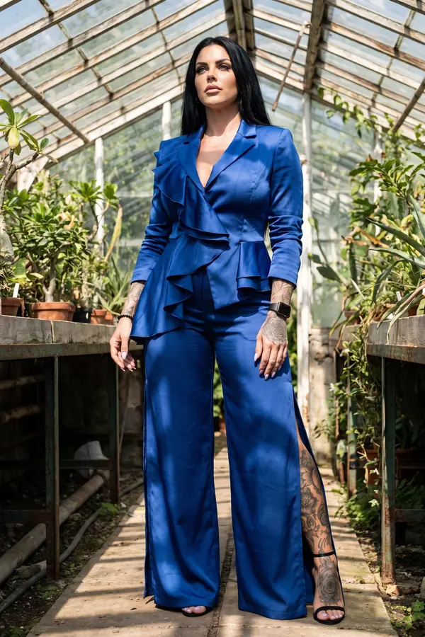 Woman in a cobalt satin ruffle blazer and matching wide-leg trousers with side slits standing in an old glasshouse