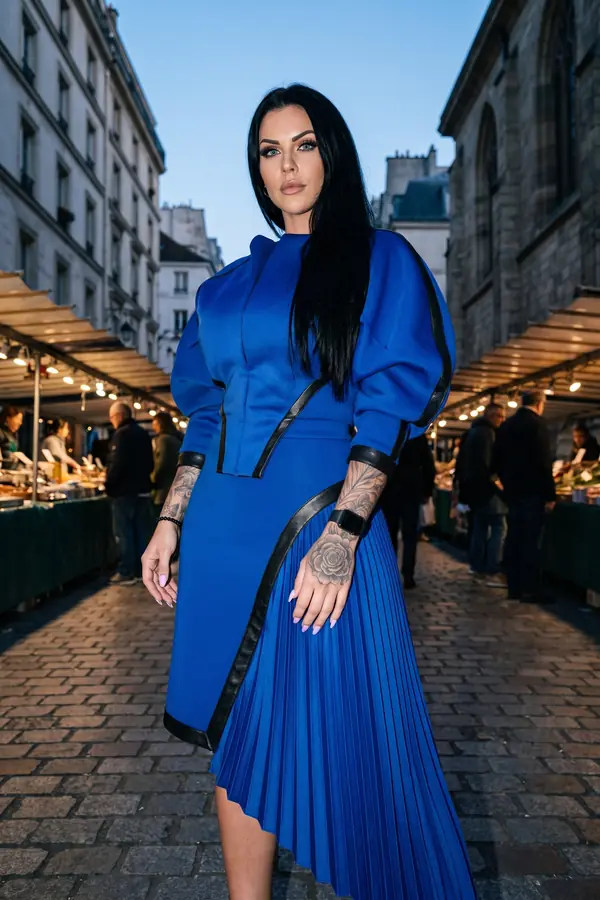 Woman in a cobalt scuba dress with black leather trim and a pleated side panel standing in a street market at dusk