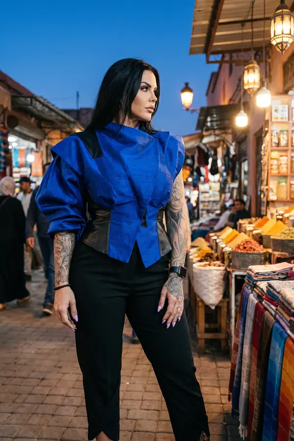 Woman in a cobalt silk shantung jacket with a black leather corset waist and cropped black trousers in a market at dusk