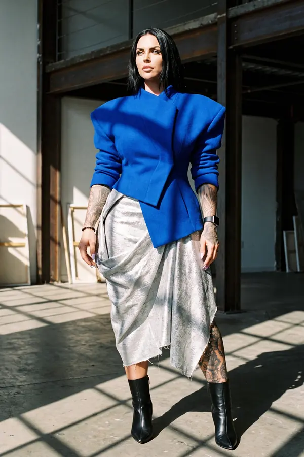 Woman in a cobalt wool jacket with sharp padded shoulders and a silver metallic draped skirt in a sunlit industrial loft