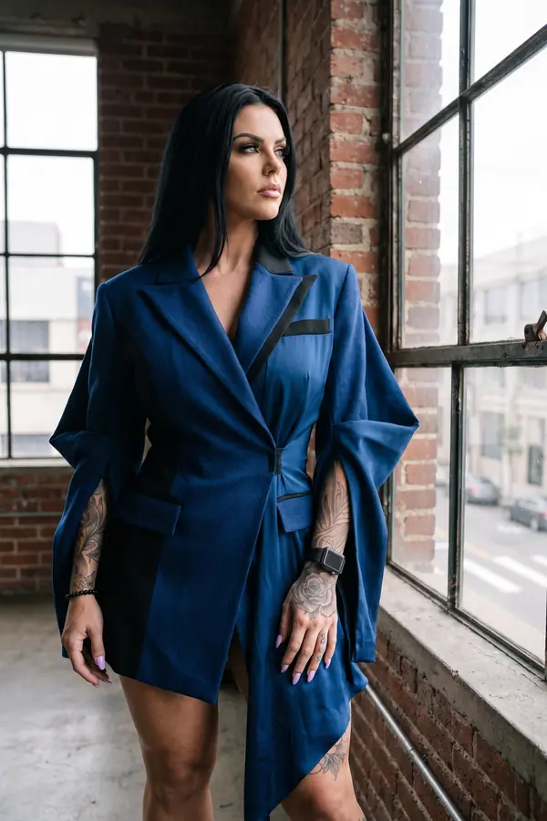 Woman in a cobalt wool blazer dress with split sleeves over a satin wrap dress by a steel window in a brick loft