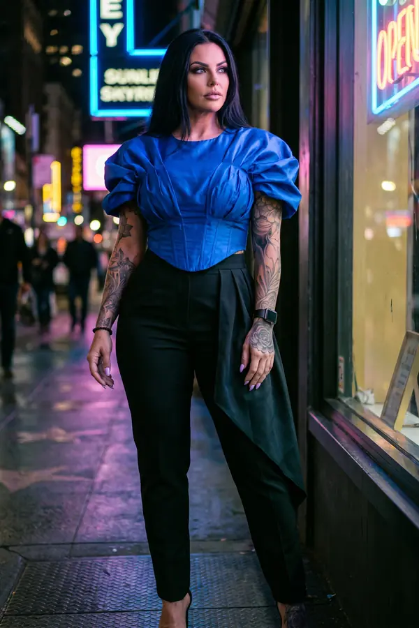 Woman in a cobalt taffeta corset top with puff sleeves and black pleated tapered trousers on a neon-lit street at night