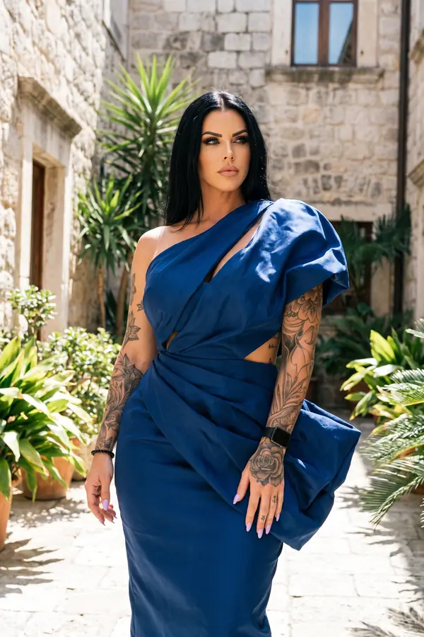 Woman in a cobalt taffeta one-shoulder dress with a sculpted draped sleeve standing in a sunlit old stone courtyard