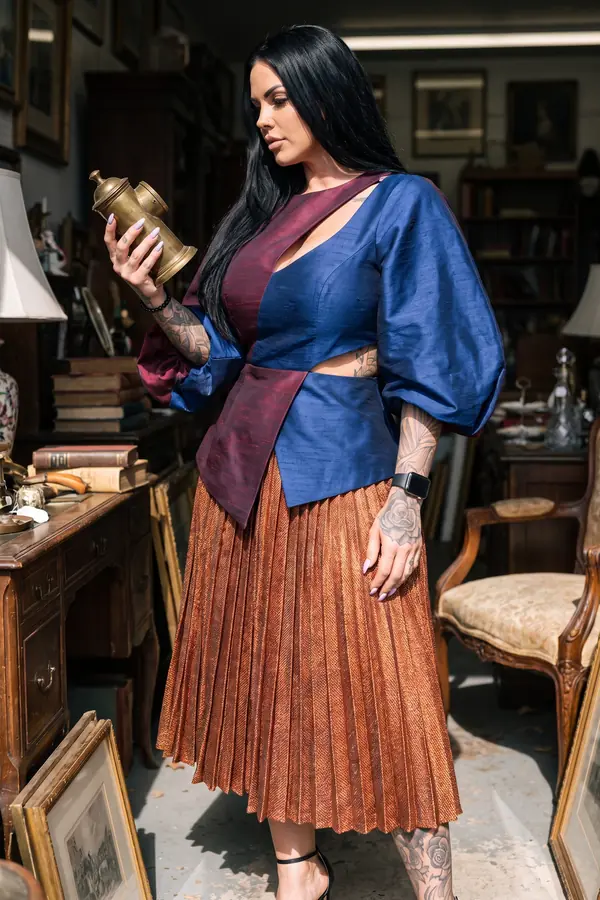Woman in a navy and burgundy silk balloon-sleeve top and copper pleated midi skirt examining a brass lamp in an antique shop