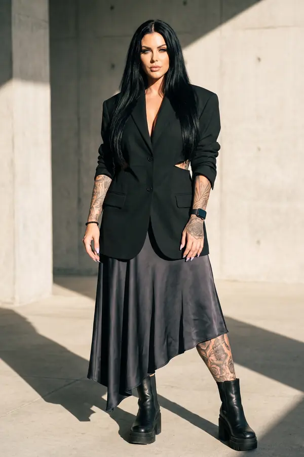 Woman in a black cut-out blazer and charcoal satin asymmetric midi skirt with chunky boots by a concrete colonnade