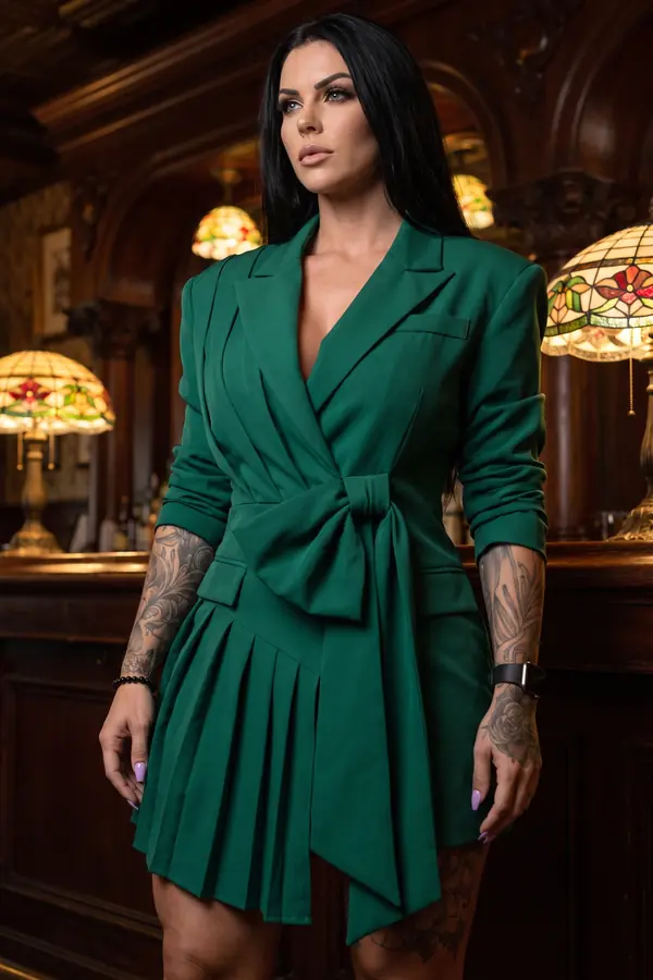 Woman in an emerald blazer dress with a waist bow and pleated panel standing at a wood-panelled bar