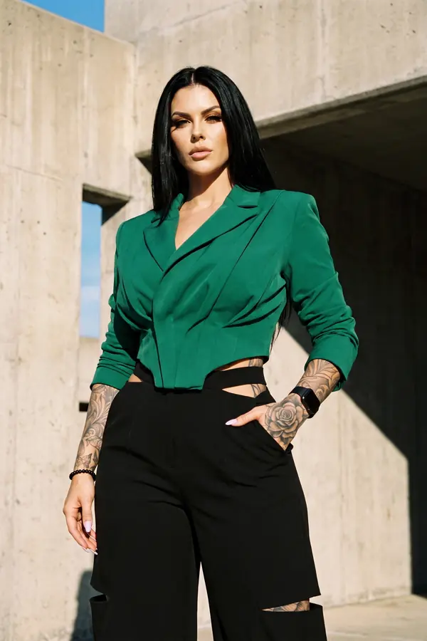 Woman in a cropped emerald blazer and black high-waisted wide-leg trousers with hip and knee cut-outs against a concrete wall