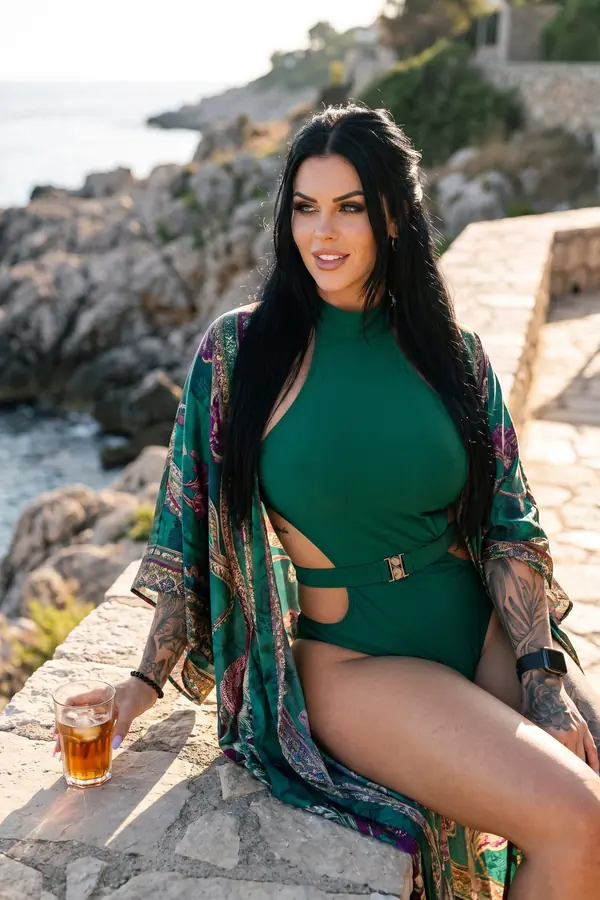 Woman in an emerald cut-out swimsuit and a green, plum and gold printed satin kimono seated on a stone wall above the sea