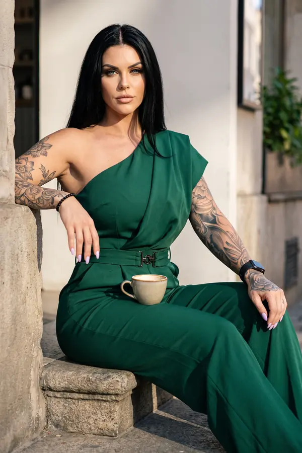 Woman in an emerald one-shoulder draped jumpsuit with a buckled belt, seated on a stone step with a coffee cup